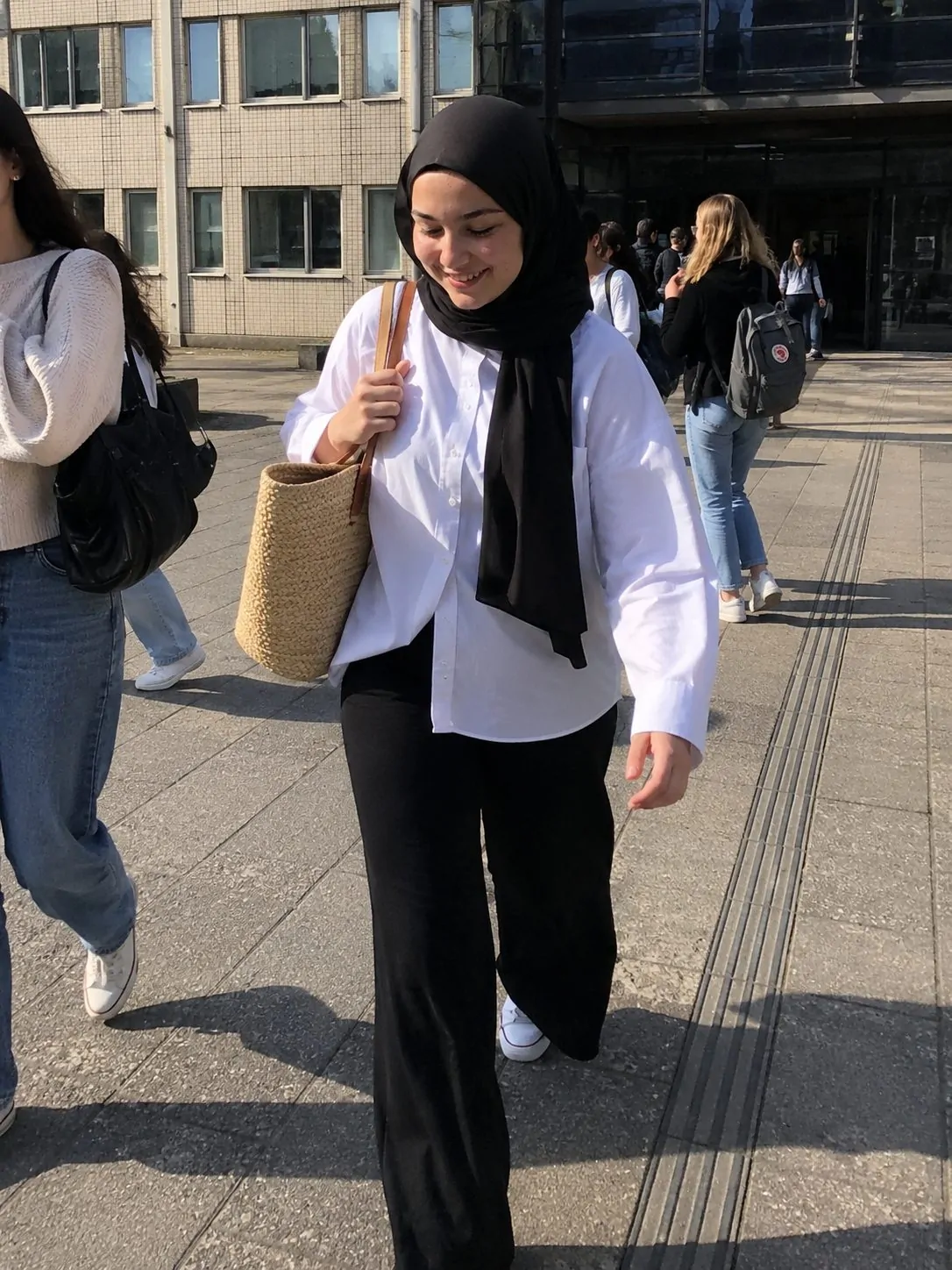 Hijabi girl in a relaxed white shirt and black bottoms with a woven bag